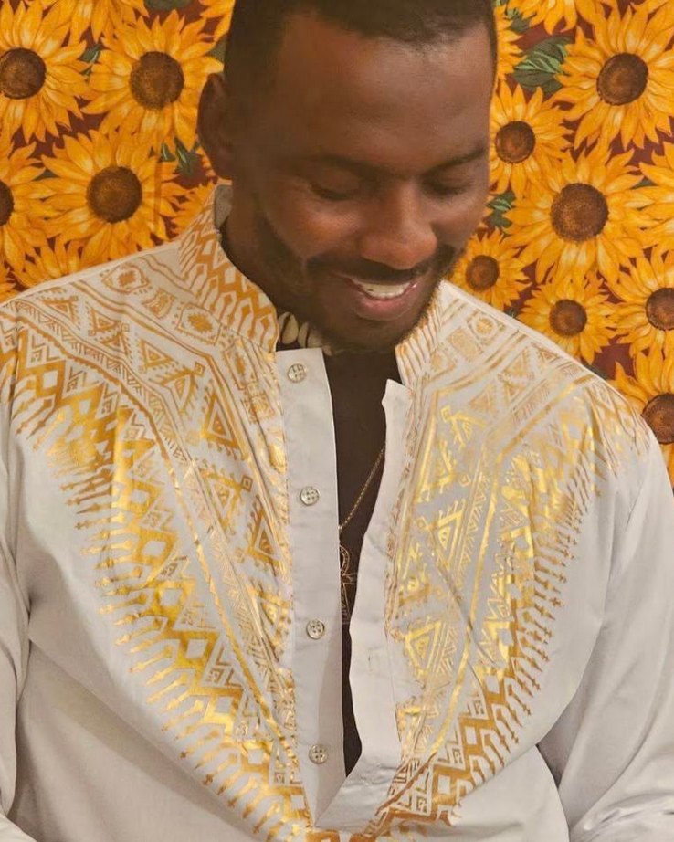 Shakespeare the Artist, smiling, in a white and gold patterned shirt in front of sunflowers