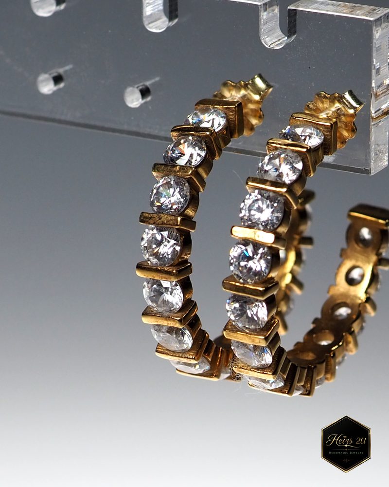 A pair of gold hoop earrings set with a row of clear stones, hanging from an acrylic stand