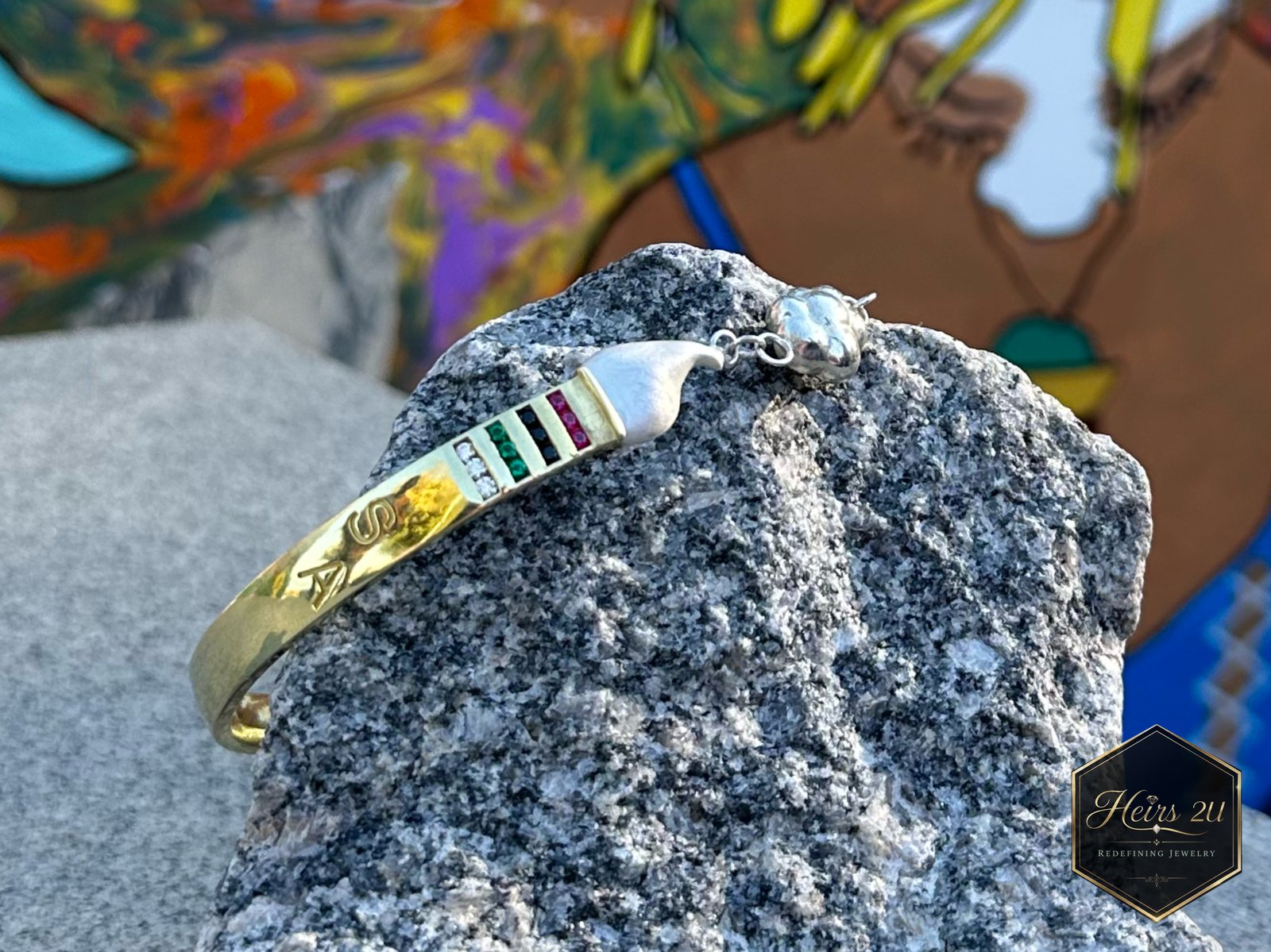 The Curved Paintbrush Bracelet by Shakespeare the Artist, a gold cuff inlaid with colour, resting on stone