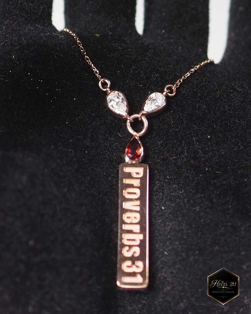 A rose-toned bar pendant engraved Proverbs 31, hung from two pear-shaped stones and a red drop, shown on black velvet
